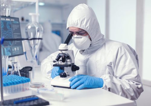 Microbiologist with protection glasses using microscope dressed ppe suit. Virolog in coverall during coronavirus outbreak conducting healthcare scientific analysis.