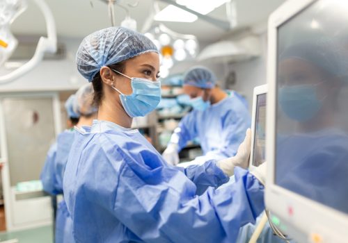 Diverse Team of Professional Surgeons Performing Invasive Surgery on a Patient in the Hospital Operating Room. Nurse Hands Out Instruments to surgeon, Anesthesiologist Monitors Vitals.